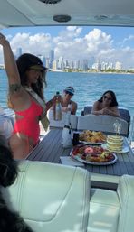 Friends celebrating on a yacht near the Miami skyline, popping champagne with cake and charcuterie on the table for a sunny waterfront party