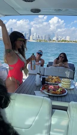 Friends celebrating on a yacht near the Miami skyline, popping champagne with cake and charcuterie on the table for a sunny waterfront party