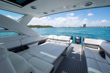Sunlit luxury yacht rear deck with white lounge seating, outboard engines, turquoise water and palm-lined coastal skyline under a blue sky.