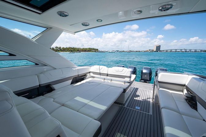 Sunlit luxury yacht rear deck with white lounge seating, outboard engines, turquoise water and palm-lined coastal skyline under a blue sky.