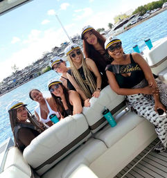 Seven women in captain hats on a sunny yacht bachelorette boat party near a marina, smiling against turquoise water and docked boats