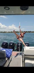 Person in a maroon bikini striking a playful pole pose on the stern of a motorboat with blue ocean and a palm-lined beach under a sunny sky.