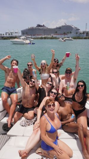 Boat party: group of friends in swimsuits cheering and holding drinks on a motorboat in turquoise harbor on a sunny day, with yachts, a large cruise ship and palm-lined shoreline in the background.
