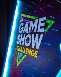 Neon-lit game show challenge sign on an indoor event stage, colorful geometric backdrop with LED accents and rows of blue pegs on an interactive prize-drop board.