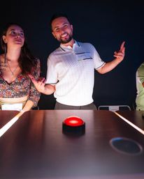 Two people at an indoor trivia game, a smiling man gesturing beside a woman as they lean over a table with a glowing red buzzer under moody lighting.
