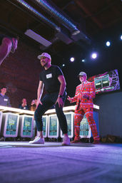 Contestant laughing and leaning forward on a lit indoor game-show stage in an industrial loft as a host in a red holiday-pattern suit gestures, illuminated podiums and a cheering audience visible.