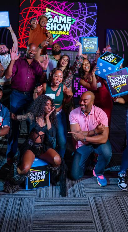 Energetic group of adults posing in a colorful game show studio, holding "The Game Show Challenge" prize boxes and giant dice in front of a neon backdrop and spinning prize wheel.