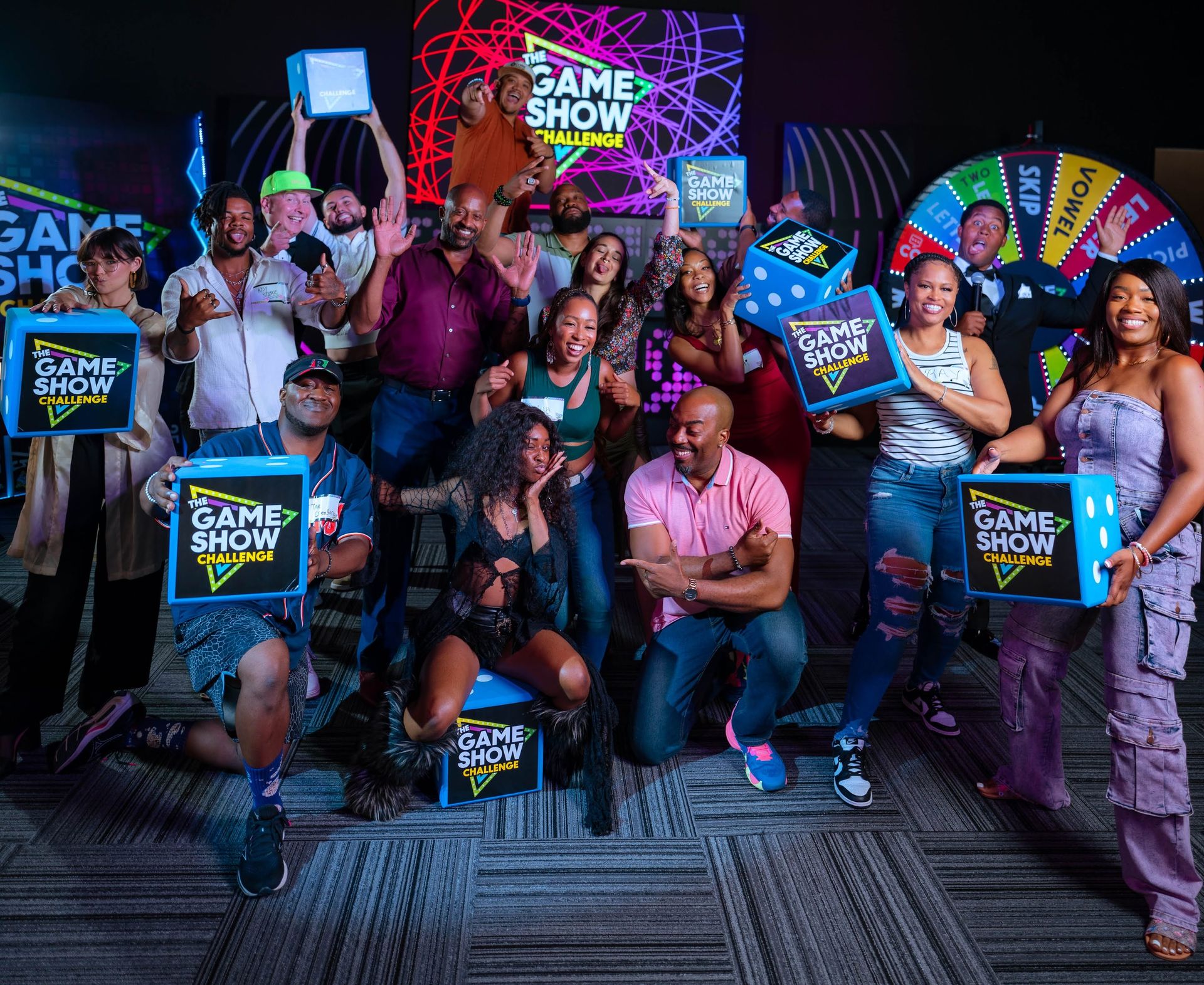 Energetic group of adults posing in a colorful game show studio, holding "The Game Show Challenge" prize boxes and giant dice in front of a neon backdrop and spinning prize wheel.