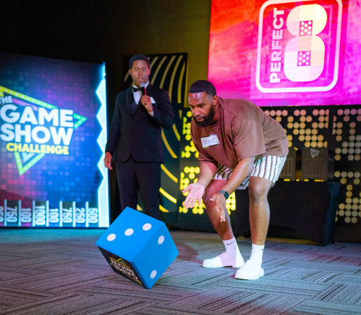 Contestant in shorts rolls an oversized blue die on a colorful indoor game-show stage while a tuxedoed host watches with a microphone