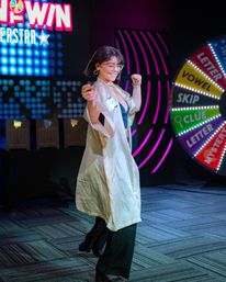 Contestant dancing and smiling on a neon-lit game-show stage, wearing a silky robe over black outfit beside a colorful spinning prize wheel labeled Vowel, Skip, Clue.