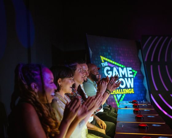 Smiling contestants cheer at buzzer stations on a colorful indoor game show stage with neon signage
