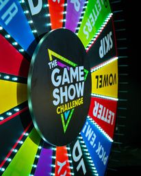 Colorful LED-lit game show prize wheel with multicolored segments and a central 'The Game Show Challenge' logo set against a dark studio backdrop