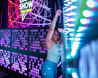 Smiling woman in a striped tank and jeans poses at a neon-lit interactive game wall in an indoor arcade, surrounded by purple and blue LED lights and a Game Show Challenge sign.
