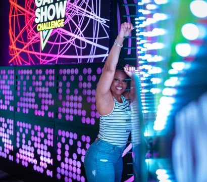 Smiling player reaches up to an interactive LED light wall in a neon-lit arcade game show attraction with colorful geometric panels.