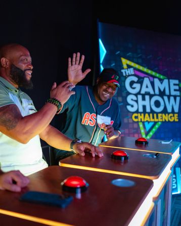 Two jubilant contestants at illuminated podiums press red buzzers during a colorful game show challenge in a studio