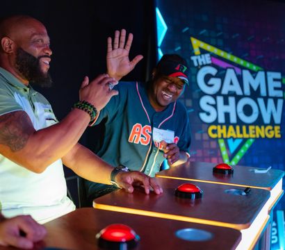 Two jubilant contestants at illuminated podiums press red buzzers during a colorful game show challenge in a studio