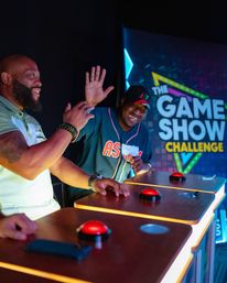 Two men laughing and high-fiving at a brightly lit game show challenge stage, pressing red buzzers on podiums with neon signage in the background.