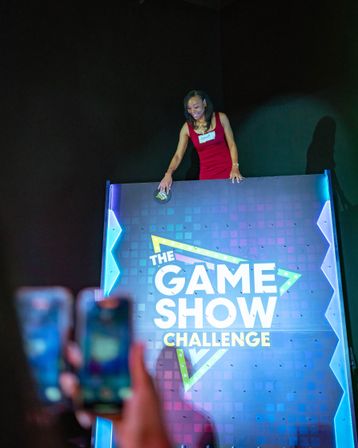 Contestant in a red dress leans over a colorful illuminated Plinko-style game board labeled THE GAME SHOW CHALLENGE while audience members capture the moment on smartphones.