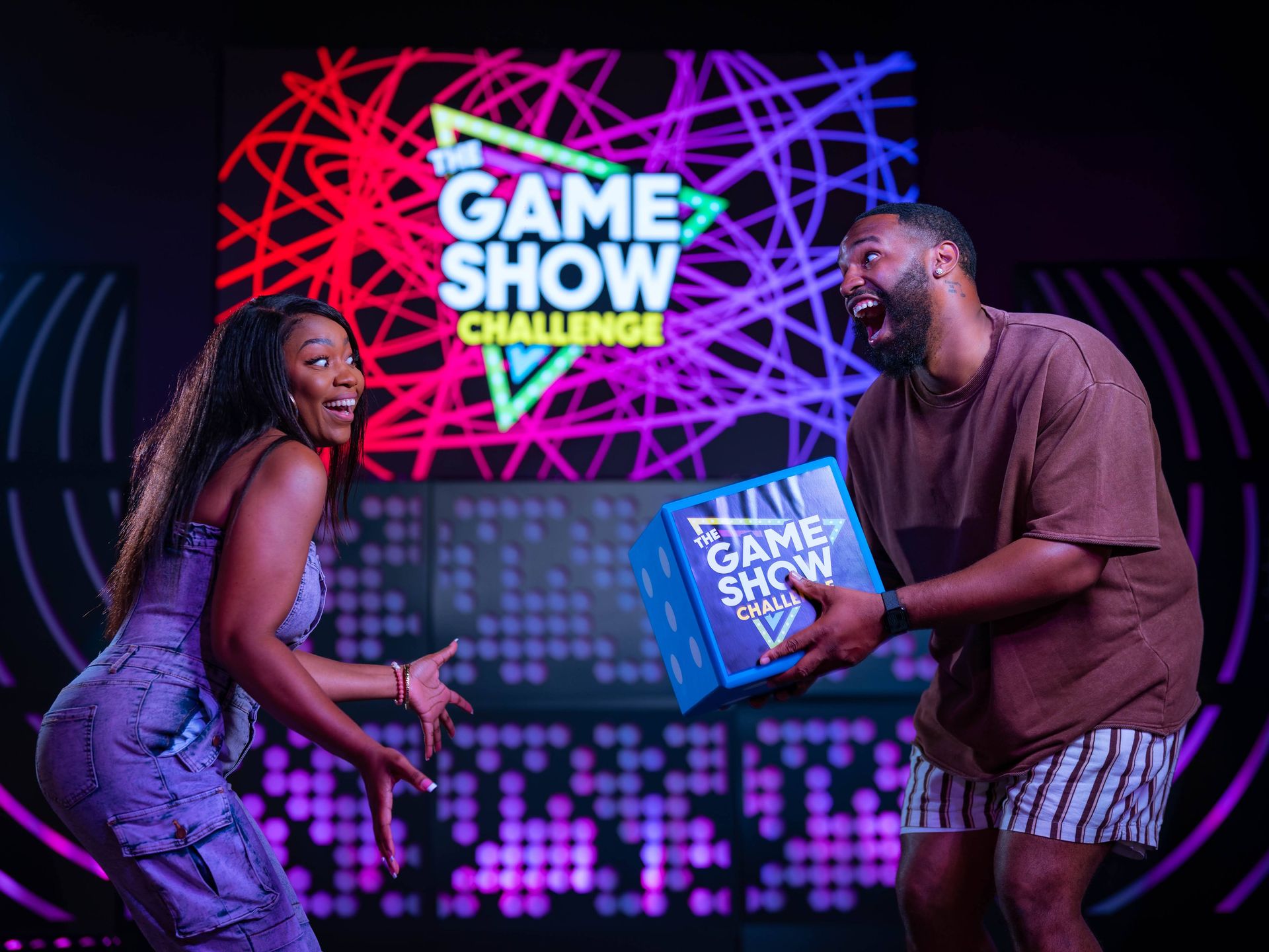 Two excited contestants laugh and reach for a blue prize box on a neon-lit indoor game show stage with colorful abstract light patterns and a large game show sign in the background.