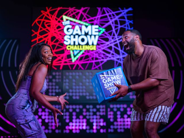 Two excited contestants laugh and reach for a blue prize box on a neon-lit indoor game show stage with colorful abstract light patterns and a large game show sign in the background.