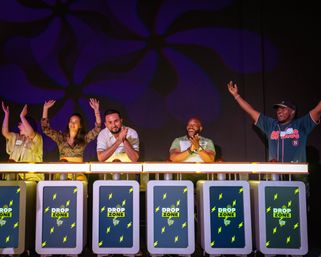 Five game-show contestants on a brightly lit studio stage cheering and clapping behind illuminated podiums with neon lightning graphics, a purple patterned backdrop, and raised hands in celebration.