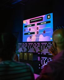 Silhouetted audience watching a colorful LED game-show screen in a neon-lit event hall, showing a multiple-choice celebrity-name matching game with a visible 36-second timer.