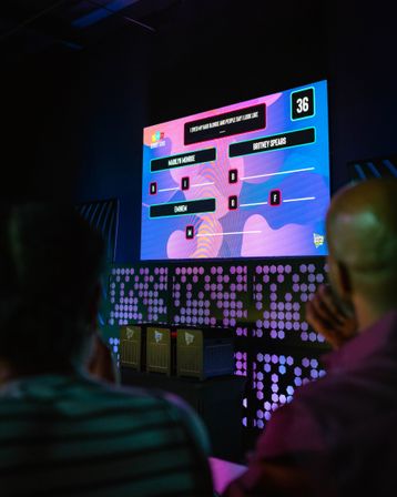 Colorful indoor game-show stage with a large LED scoreboard showing trivia answers and letters, purple-blue stage lighting and audience silhouettes in the foreground