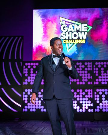 Game show host in a black tuxedo holding a microphone on a colorful LED stage with a 'The Game Show Challenge' logo and purple event lighting