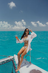 Woman in white bikini and open shirt wearing sunglasses, sipping wine on the bow of a catamaran over clear turquoise tropical ocean under a sunny blue sky with fluffy clouds.