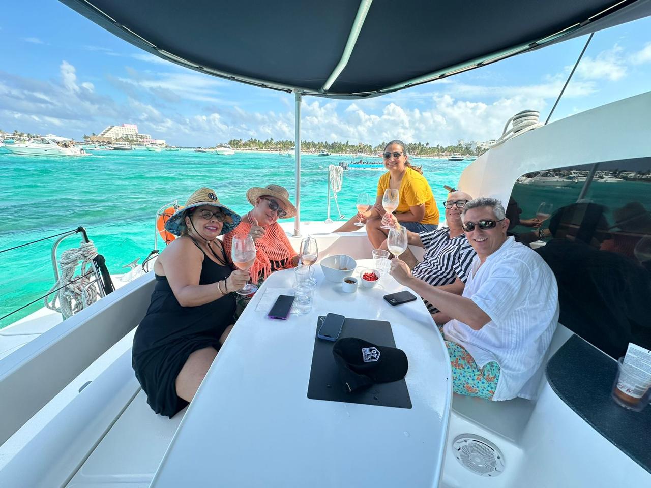 Five adults smiling and toasting with wine on the shaded deck of a white catamaran over turquoise tropical water, with palm-lined shoreline and boats in the background.