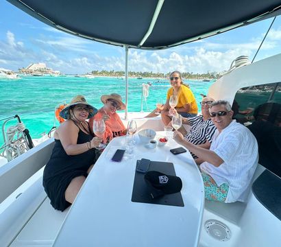 Five adults smiling and toasting with wine on the shaded deck of a white catamaran over turquoise tropical water, with palm-lined shoreline and boats in the background.