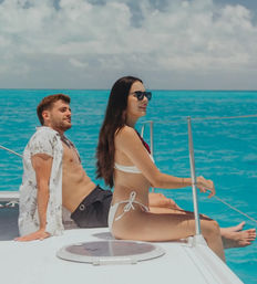 Couple relaxing on a white boat deck over turquoise tropical sea — woman in a white bikini and sunglasses smiling at the horizon, man reclining shirt open under a sunny, cloud-dotted sky, enjoying a sailing vacation.