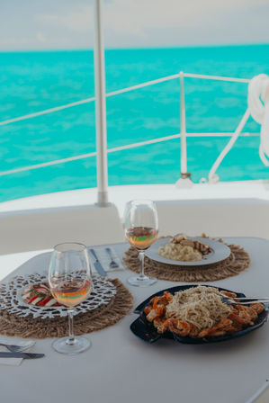 Sunlit boat-deck dining with two glasses of rosé and plates of shrimp pasta, salad and sides on a white table overlooking turquoise ocean