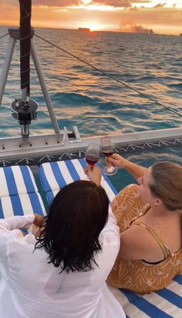 Two people toasting red wine on a sailboat trampoline with striped cushions, watching a golden ocean sunset on a sunset sail — relaxing coastal vacation vibe.