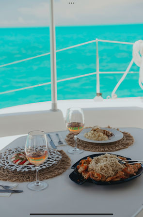 Sunlit sailboat lunch: two glasses of rosé and a seafood pasta with sides on a white table, placemats and bright turquoise tropical sea in the background.