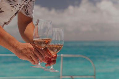 Two hands holding rosé wine glasses aboard a boat with turquoise ocean and cloudy sky in the background