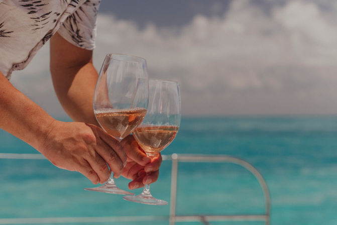 Two hands holding rosé wine glasses aboard a boat with turquoise ocean and cloudy sky in the background