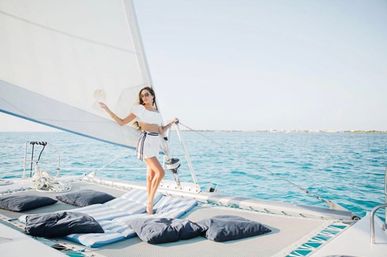 Woman in a white summer outfit standing on a catamaran trampoline with blue cushions, large white sail and turquoise sea under a clear sky — sunny sailing escape.
