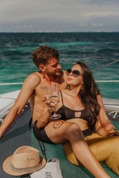 Couple lounging on a sailboat over turquoise tropical ocean — woman in black bikini and sunglasses sipping white wine while a shirtless man leans in, sun hat and beach tote on the deck.