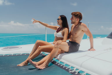 Young couple in swimwear relaxing on a sailing catamaran trampoline, woman pointing toward the turquoise tropical sea under a clear blue sky