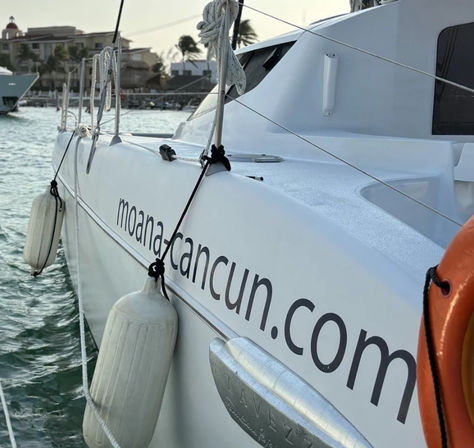 White catamaran docked in a Cancun marina, showing hanging white fenders, orange lifebuoy, calm turquoise water and palm-tree waterfront buildings in the background.