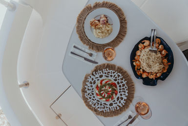 Overhead view of al fresco yacht dining: white table set with two glasses of rosé and three dishes — a caprese-style tomato and mozzarella salad with pesto, a plate of pasta surrounded by sautéed shrimp, and sliced grilled protein on creamy mashed potatoes.