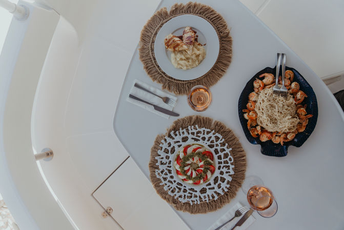 Overhead view of al fresco yacht dining: white table set with two glasses of rosé and three dishes — a caprese-style tomato and mozzarella salad with pesto, a plate of pasta surrounded by sautéed shrimp, and sliced grilled protein on creamy mashed potatoes.