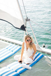 Sunlit woman in a white jumpsuit and sunglasses relaxing on blue-and-white striped cushions on a catamaran net, smiling with a phone and water bottle, sail rigging and calm turquoise sea in the background.