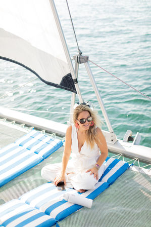 Sunlit woman in a white jumpsuit and sunglasses relaxing on blue-and-white striped cushions on a catamaran net, smiling with a phone and water bottle, sail rigging and calm turquoise sea in the background.