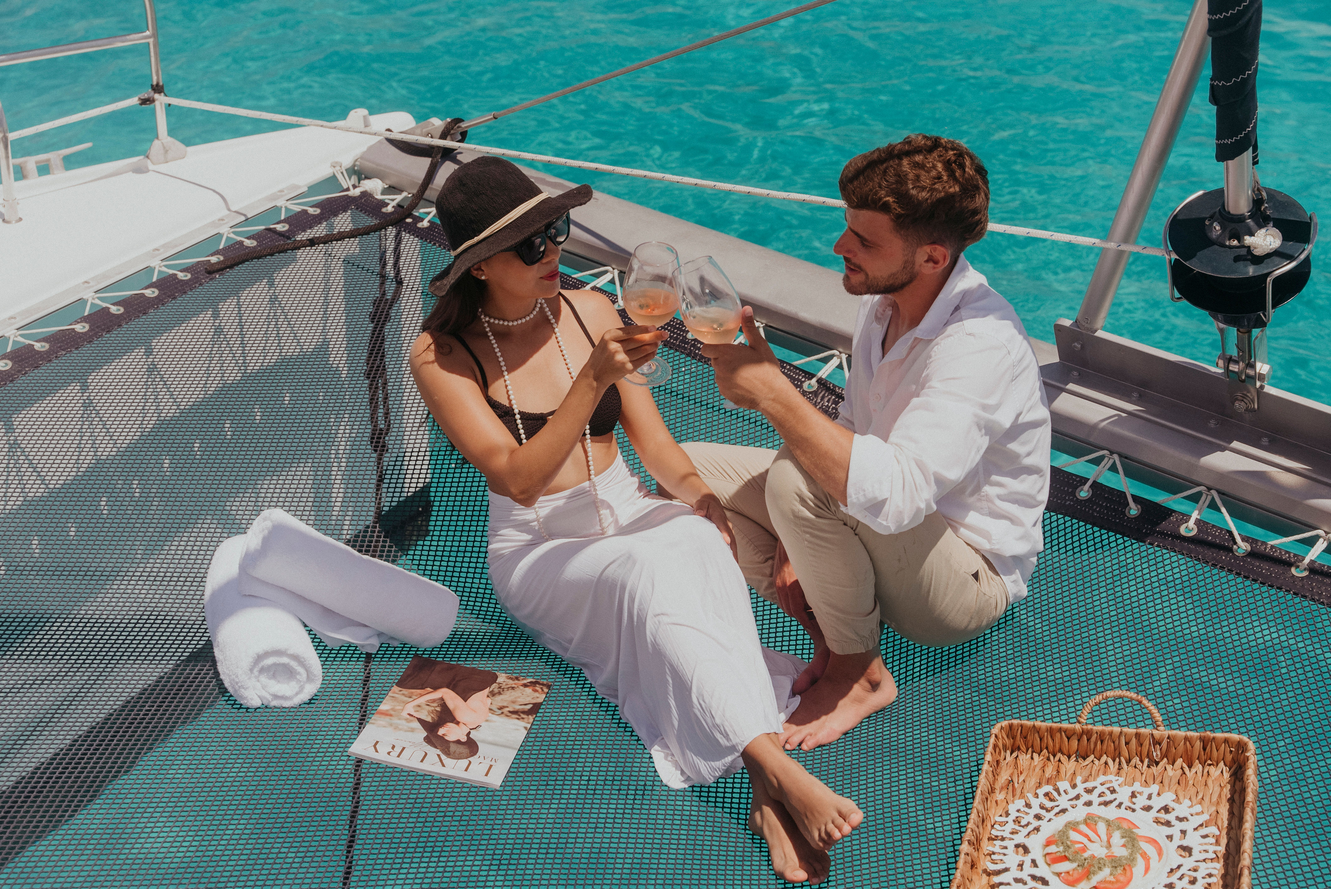 Couple toasting rosé on a catamaran trampoline over turquoise tropical sea—woman in sunhat and white skirt and man in a linen shirt, with rolled towel, magazine, and picnic basket nearby.