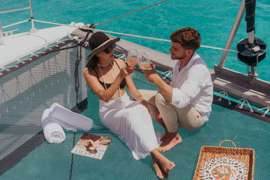 Couple toasting rosé on a catamaran trampoline over turquoise tropical sea—woman in sunhat and white skirt and man in a linen shirt, with rolled towel, magazine, and picnic basket nearby.
