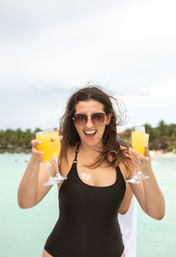 Smiling woman in a black swimsuit and sunglasses holding four glasses of orange mimosas on a tropical beach with turquoise water and palm trees in the background.