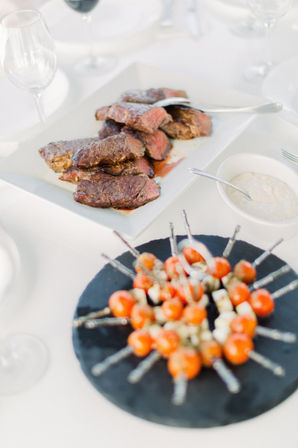Sliced medium-rare steak on a white platter with cherry tomato and mozzarella skewers on a round slate board, dipping sauce and wine glasses on a white tablecloth