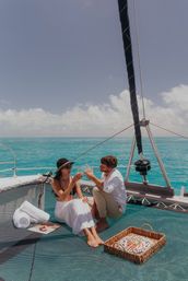 Couple toasting with wine on a catamaran trampoline above clear turquoise tropical waters, sunlit sky, picnic basket and rolled towels for a relaxed sailing getaway.